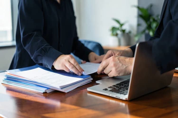 Uma pessoa organizando papéis em uma mesa com um laptop aberto ao lado, em um escritório moderno.